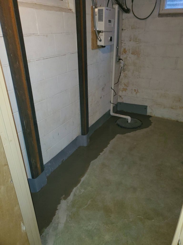 Basement corner with steel wall braces installed on concrete block walls, perimeter trench filled with new concrete, and sump pump discharge system.