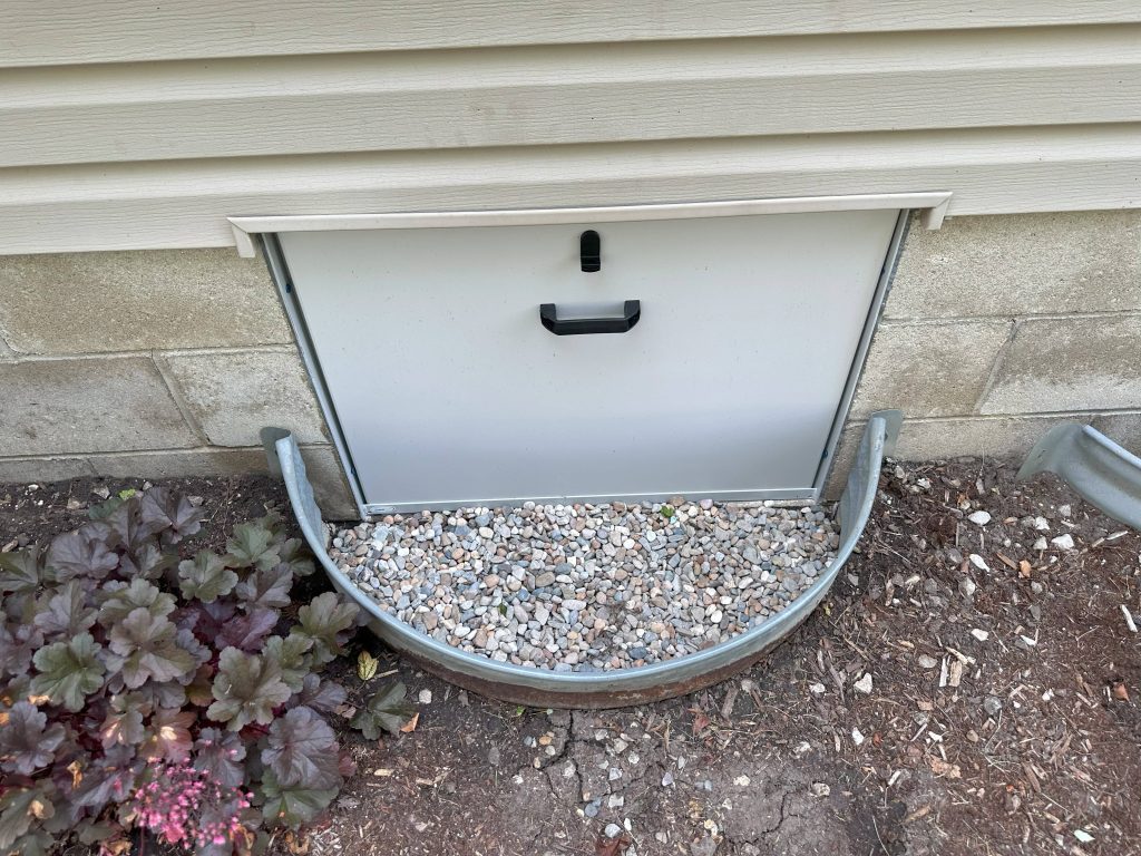 Exterior crawl space entry door with metal well and gravel base set against a home’s foundation wall.