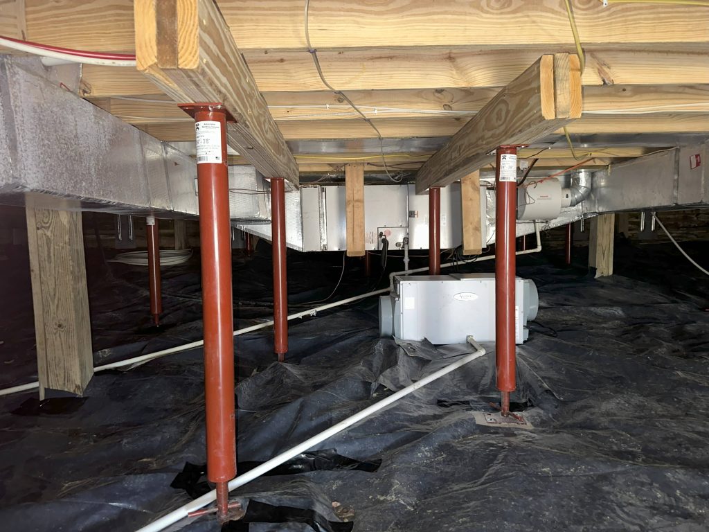 Crawl space with red steel floor support jacks installed under wooden beams, along with vapor barrier and HVAC ductwork.