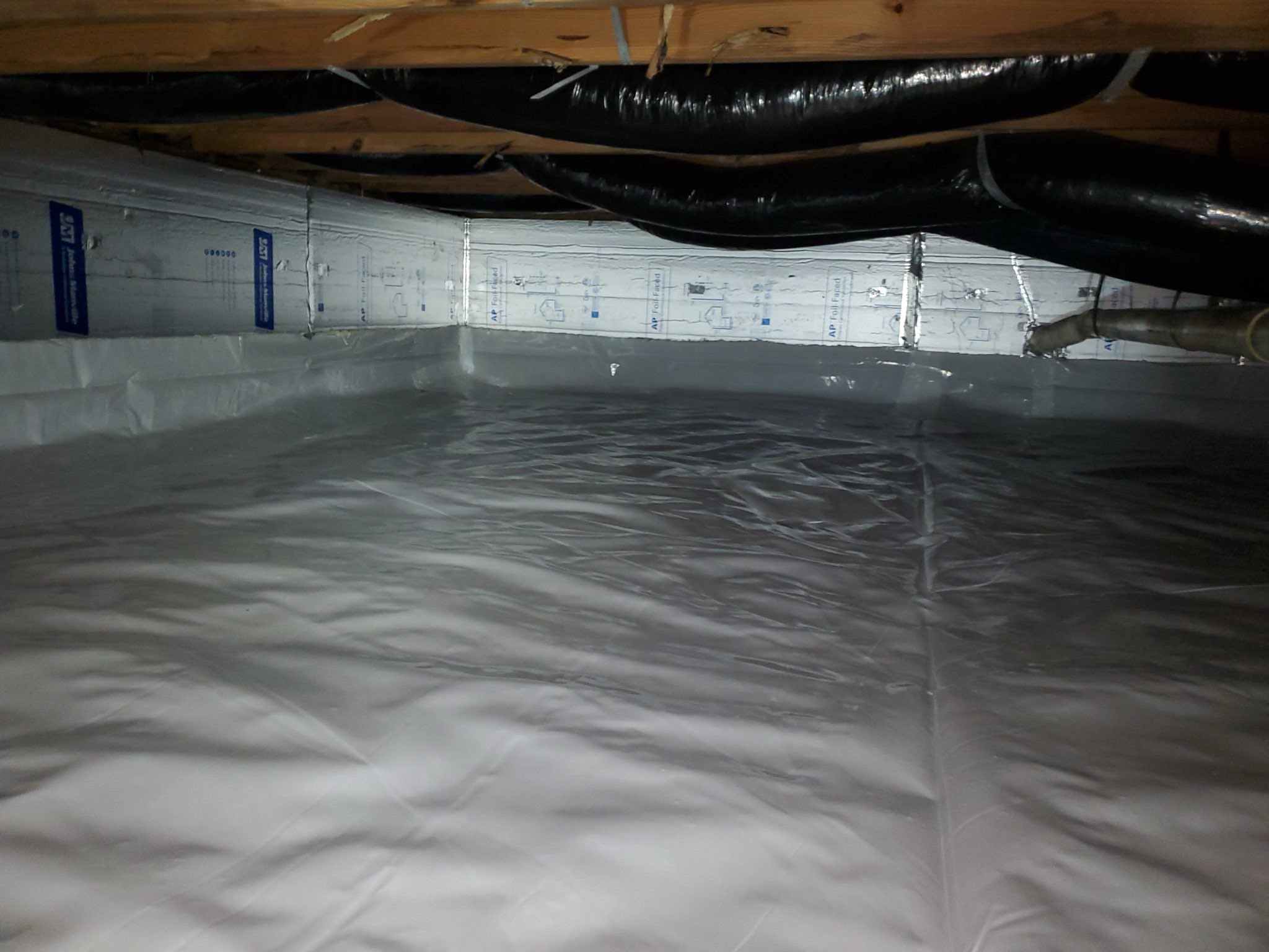 Crawlspace with installed insulation and white vapor barrier lining the floor and walls, with overhead ductwork visible